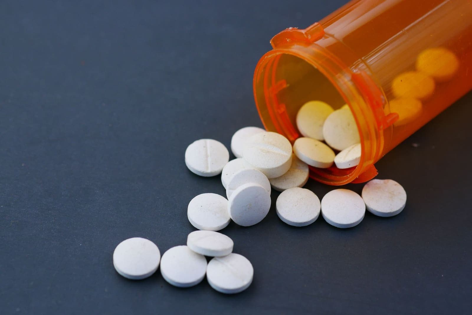 White tablets spilling out of an orange prescription bottle onto a dark surface