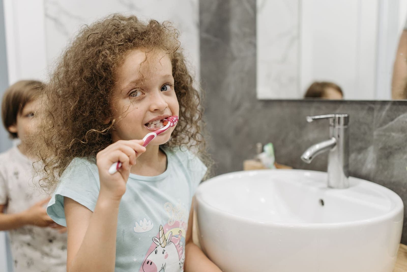 A young girl with curly hair brushes her teeth at a bathroom sink while another child stands behind her