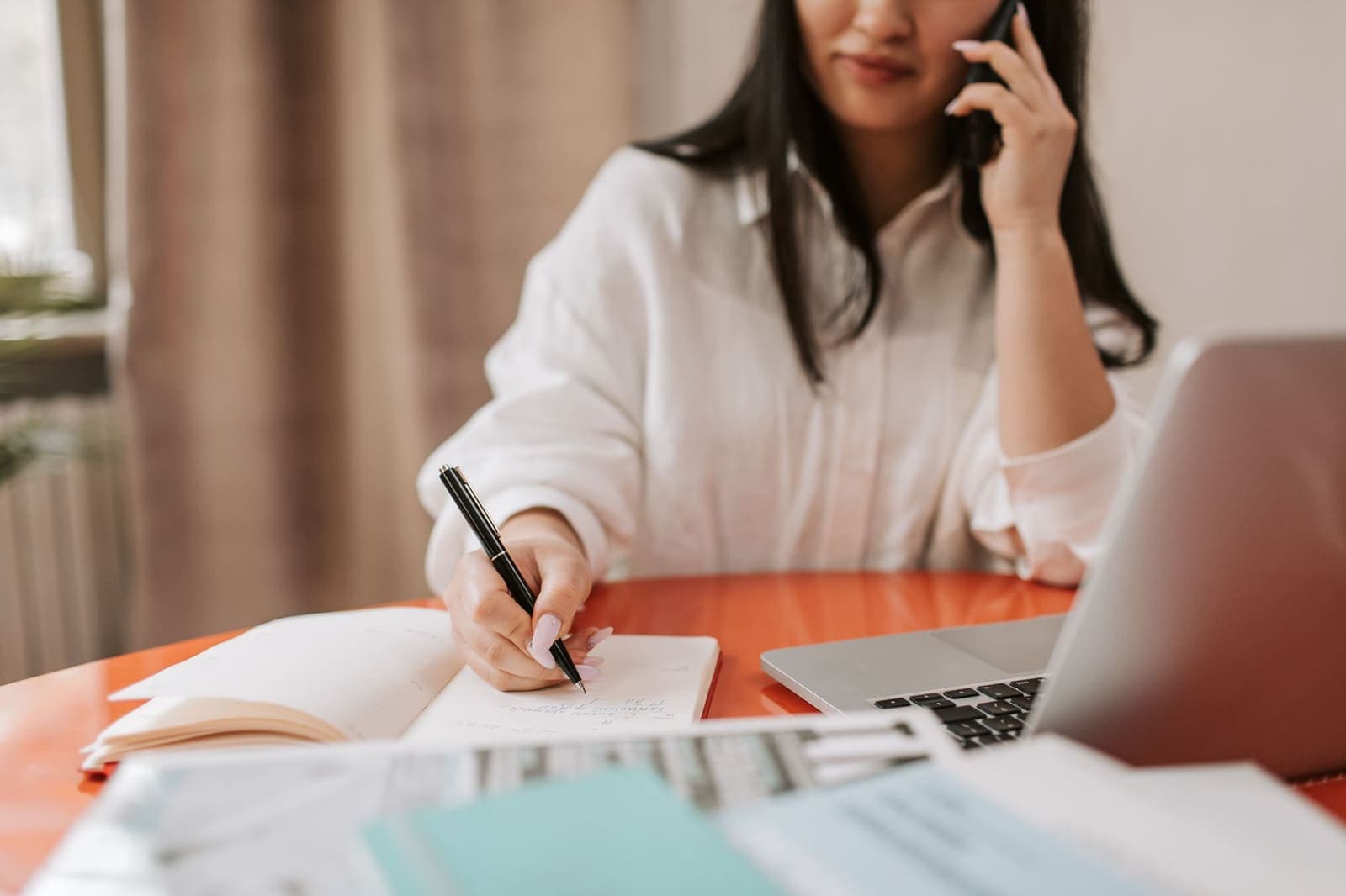 A woman on a phone call writes notes in a notebook at a desk beside a laptop