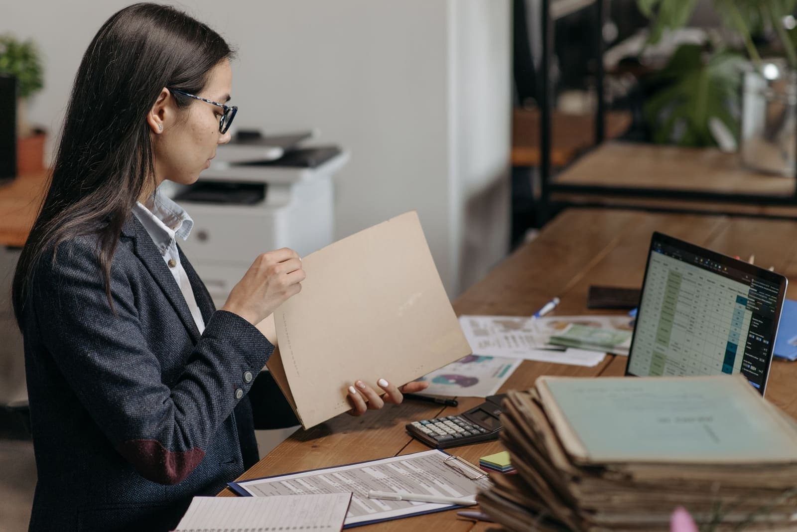 A practice manager in a dark blazer looks through a manila folder at a desk with a laptop spreadsheet, a calculator and a stack of files