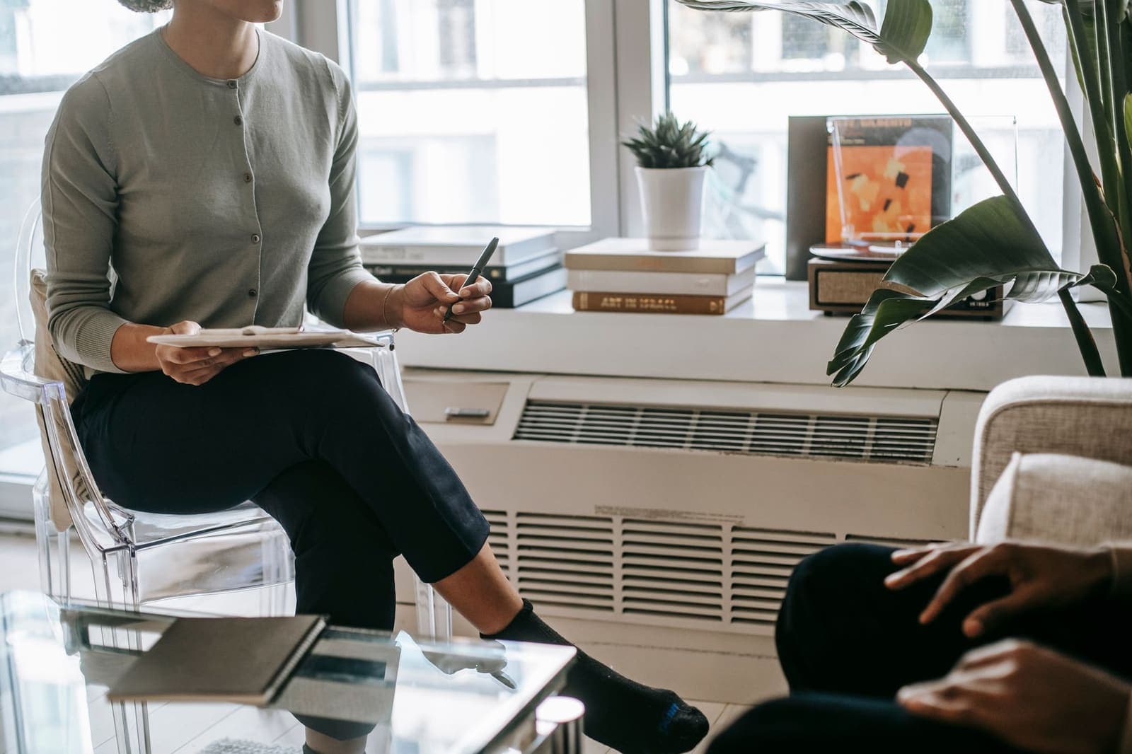 A therapist in a grey cardigan sits with a clipboard and pen in a bright office, facing a client on a sofa