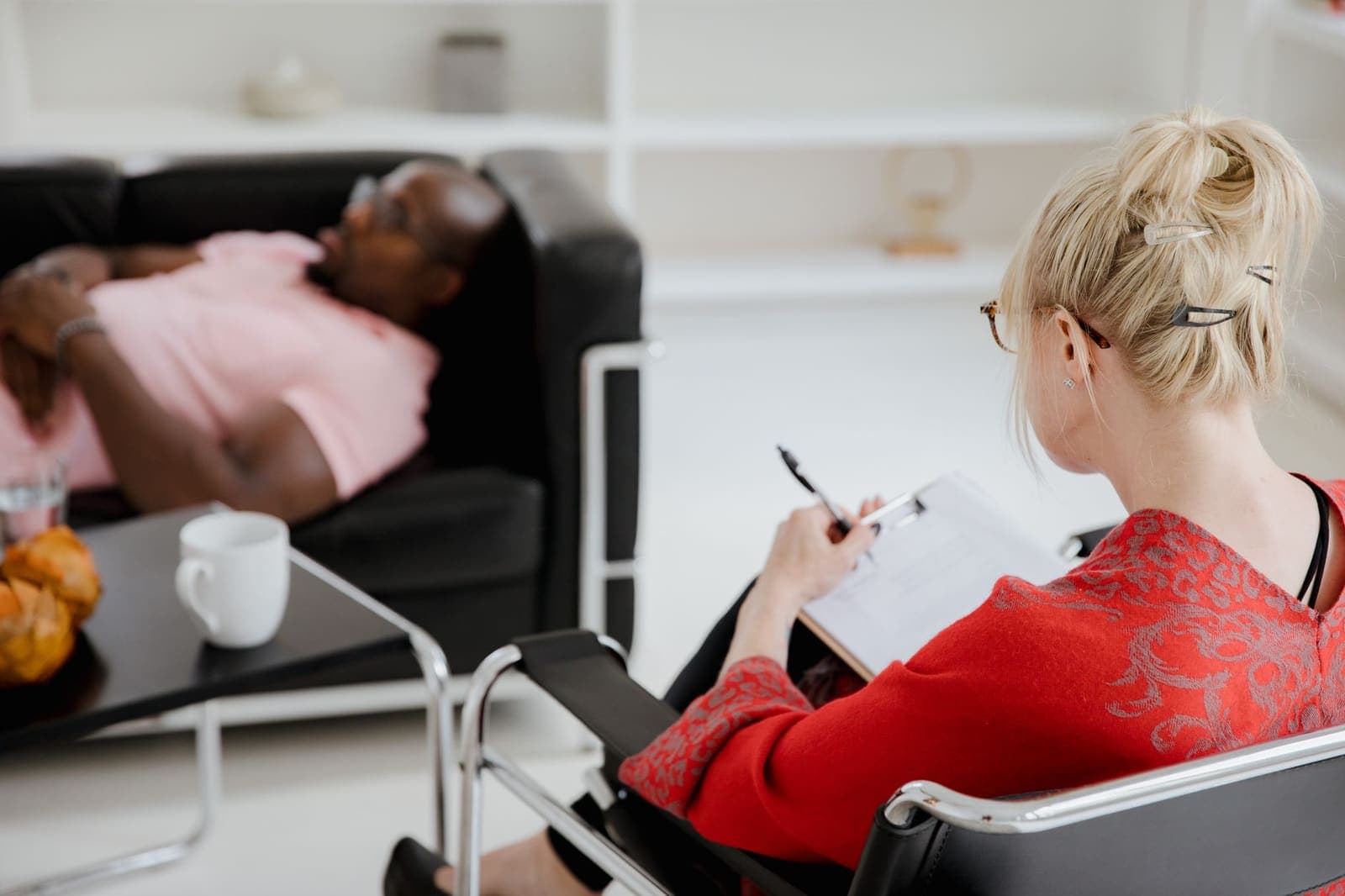 A therapist in a red top writes on a clipboard while a client lies back on a black leather sofa