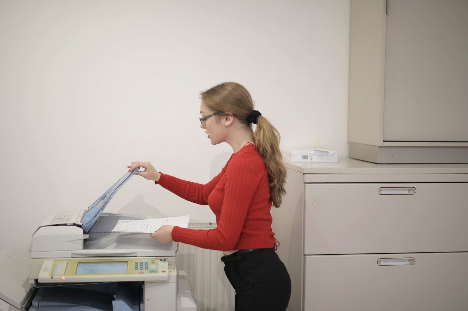A woman in a red sweater feeding pages into an office copier beside a grey filing cabinet