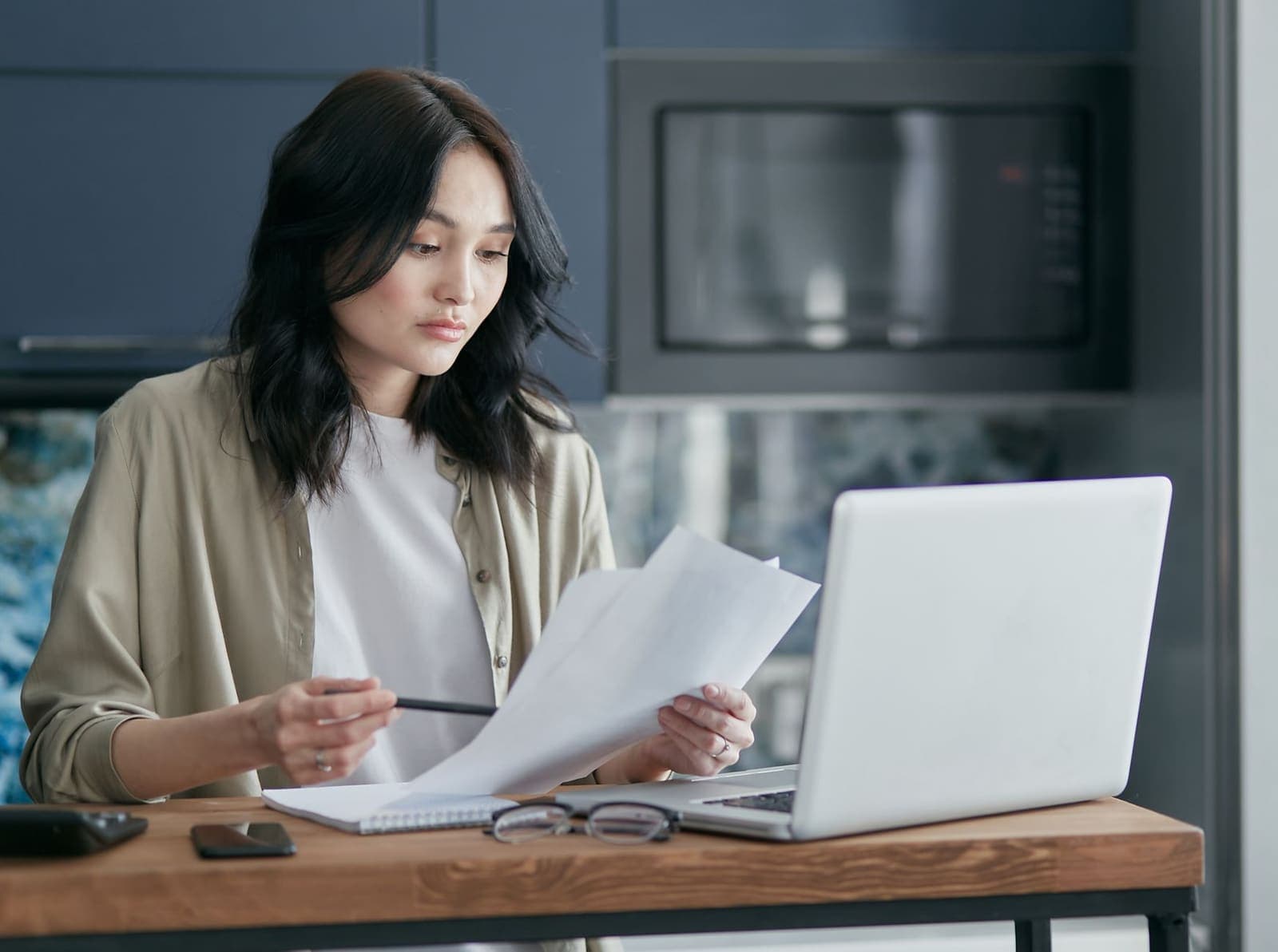 A woman at a table at home reading printed pages beside an open laptop, a notebook and her glasses
