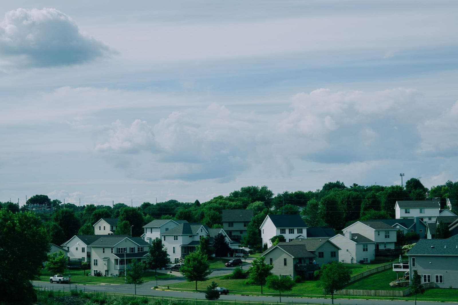 A Midwest subdivision of two-story houses with gray and white siding on a green hillside under a cloudy summer sky