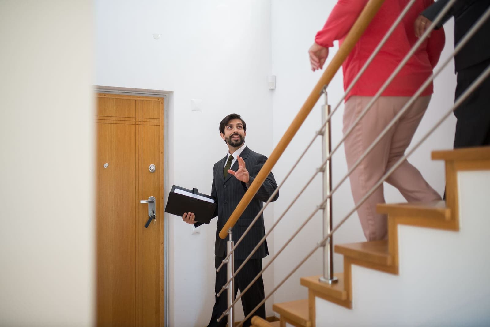 A real estate agent in a dark suit holding a folder gestures up a staircase as two clients climb ahead of him