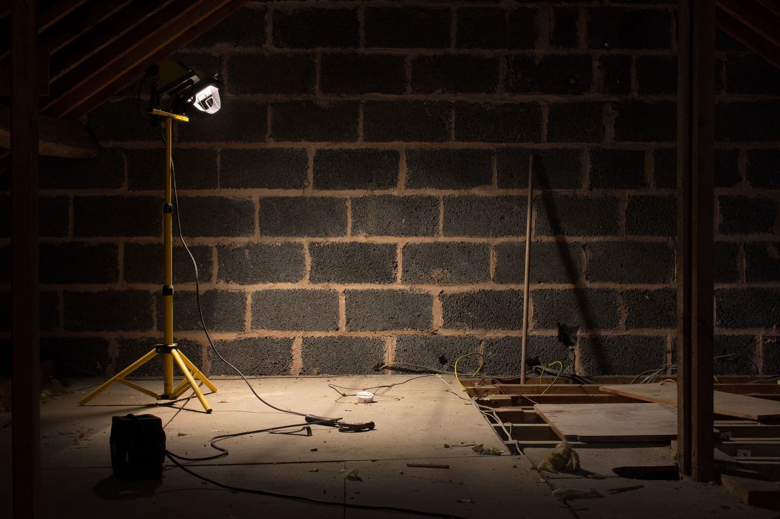 A dim, unfinished attic lit by a single work light on a yellow tripod, with loose wiring and open floor joists