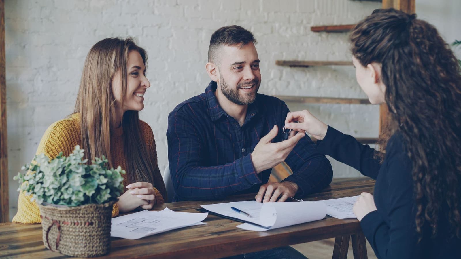 A smiling young couple at a wooden table receiving a set of house keys from a woman across the table