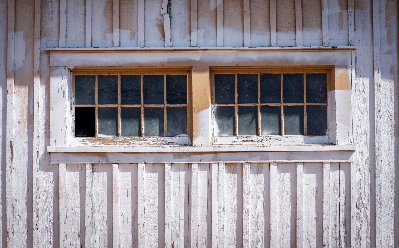 Two old multi-pane windows in a white board-and-batten wall with peeling paint and one missing pane