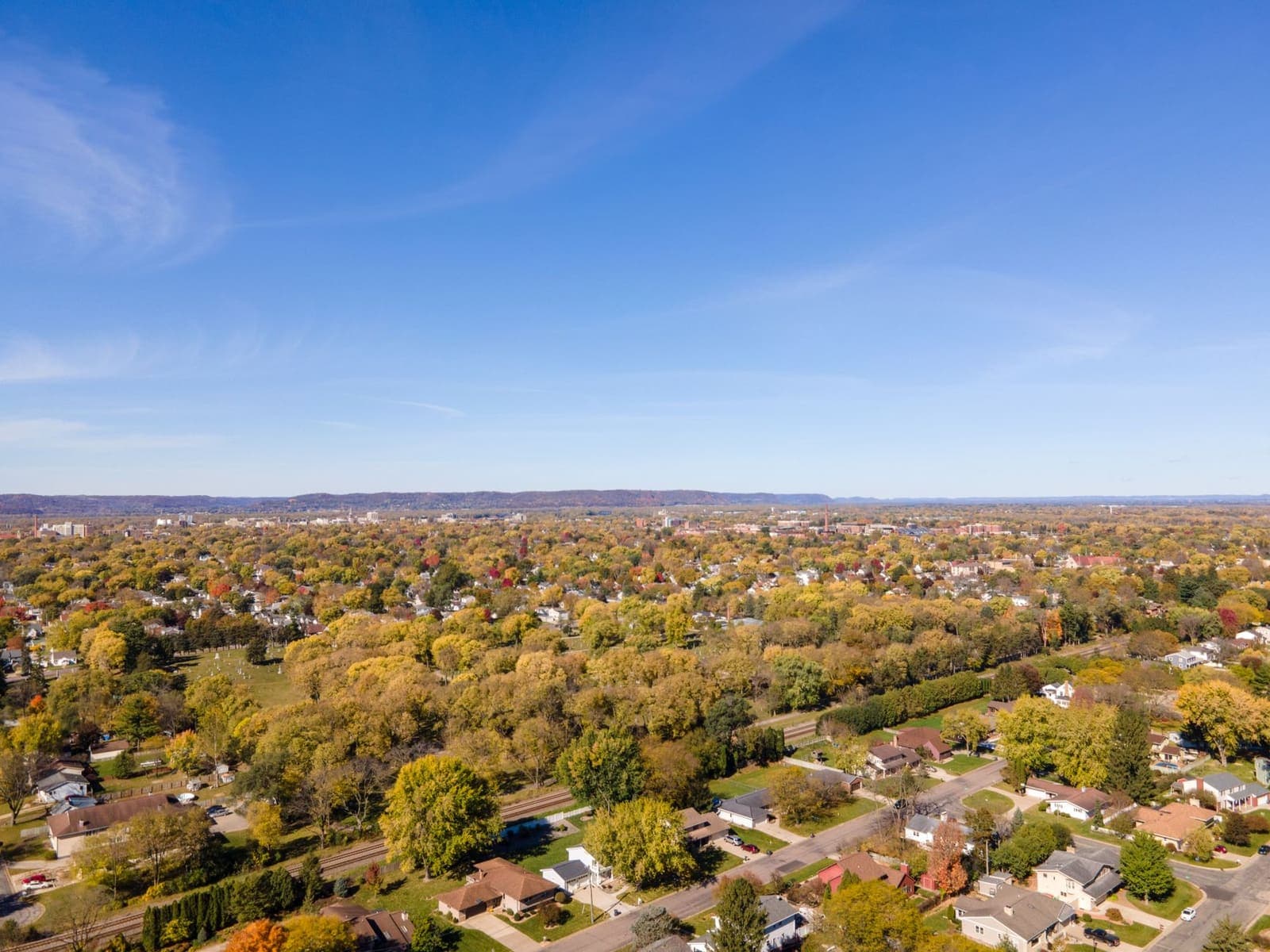 An aerial view of a Midwest town in autumn, with houses and streets under yellow and orange trees and a line of low hills on the horizon