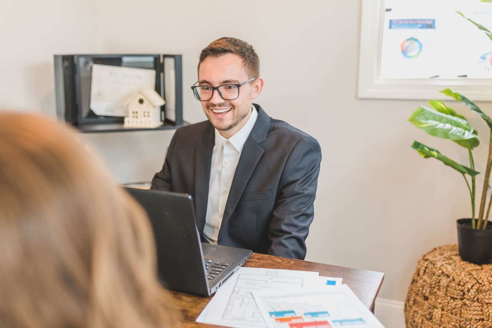 A smiling agent in a dark jacket at a desk with a laptop and printed floor plans, facing a client in the foreground