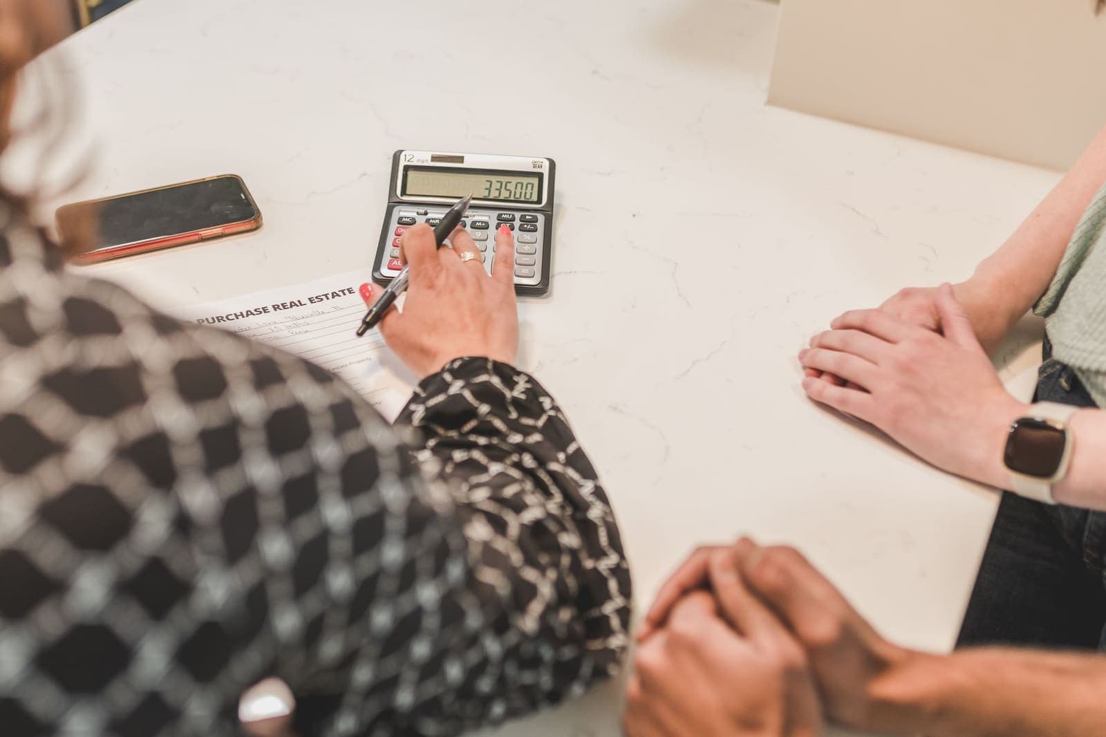 A real estate agent taps figures into a calculator beside a purchase agreement while two clients wait across the table