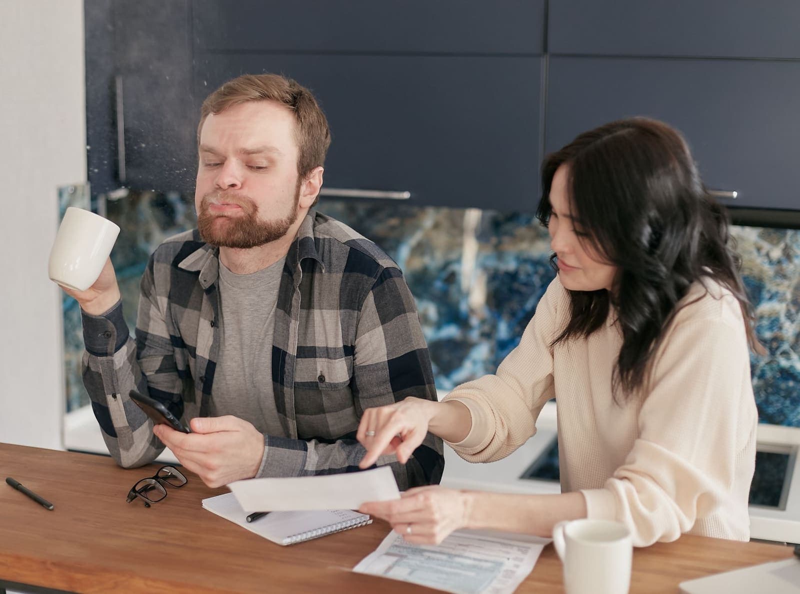 A couple at a kitchen counter going through paperwork, one holding a mug and a phone, the other pointing at a form
