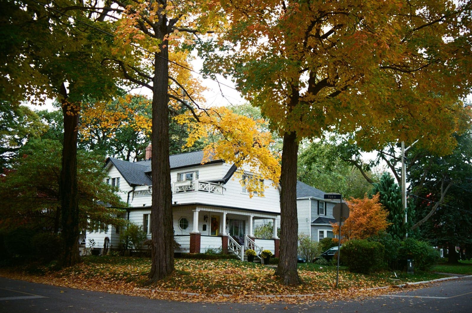 A white two-storey house with a deep front porch on a corner lot, under maple trees turning yellow and orange