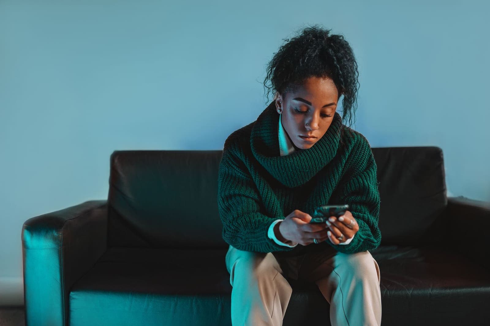 A woman in a green sweater sitting on a black sofa, reading her phone