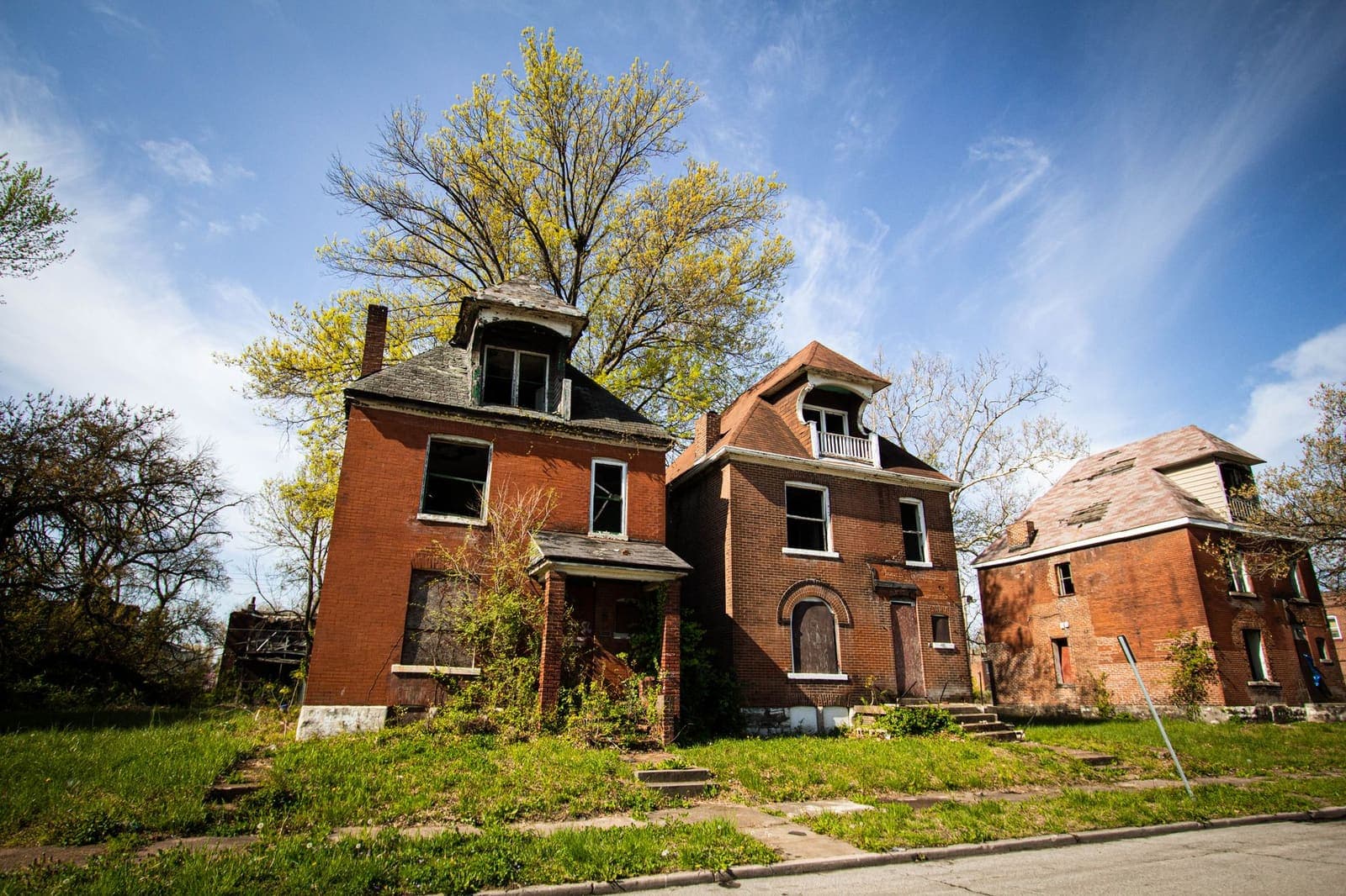 Three vacant two-storey brick houses with boarded and broken windows on an overgrown city block under a blue sky
