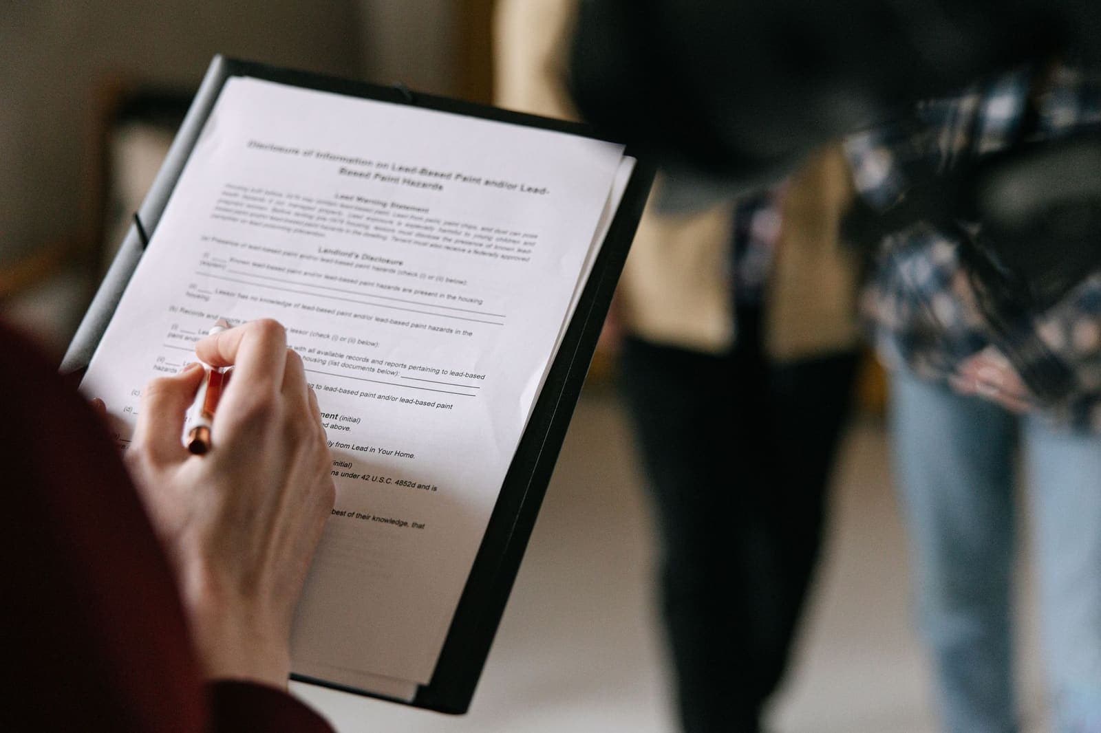A person filling in a lead-based paint disclosure form on a clipboard while two people stand nearby