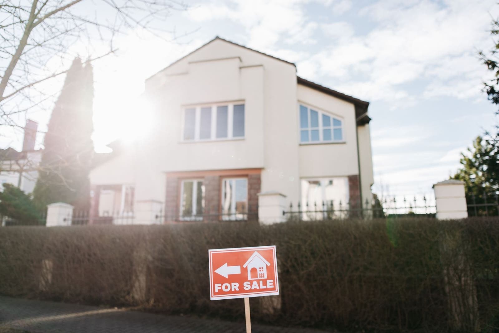 A red for sale sign on a post in front of a hedge and a cream two-storey house