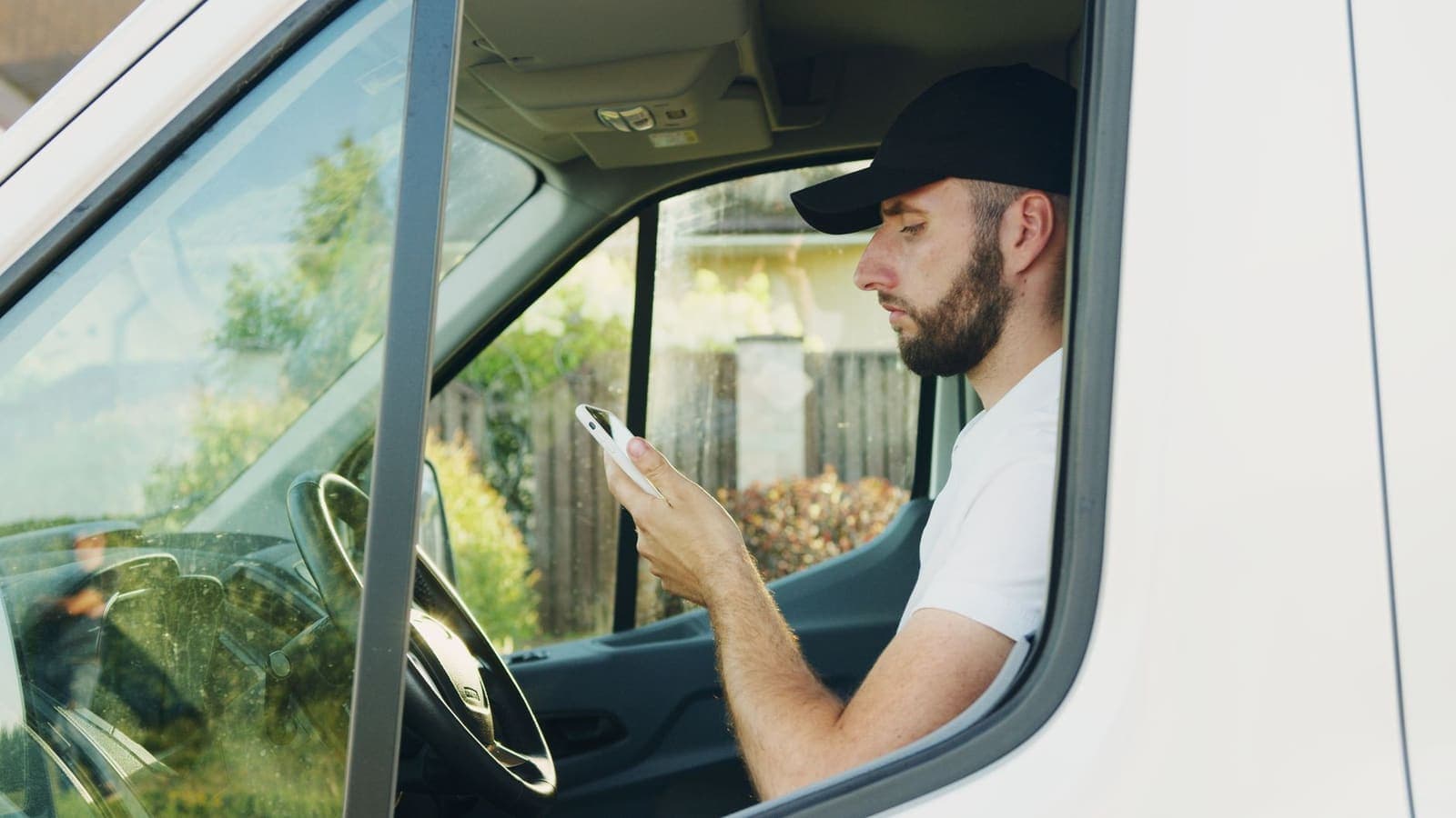 A man in a black cap sitting in the driver's seat of a white van, reading his phone
