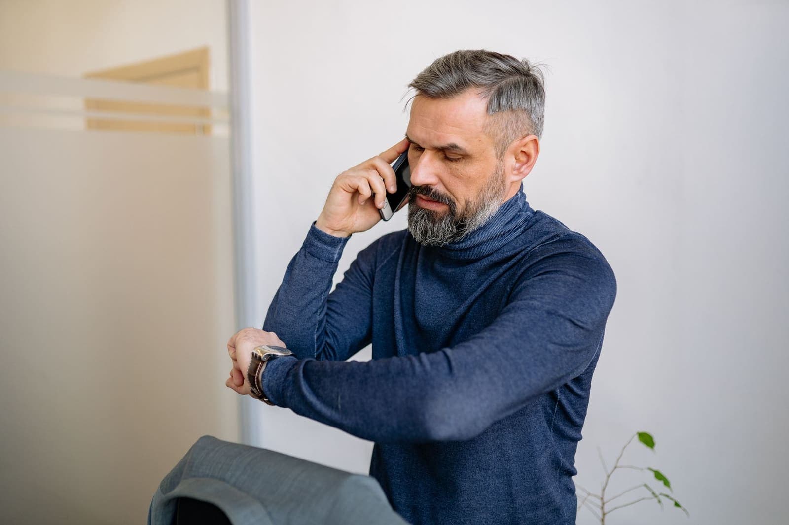 A grey-bearded man in a navy roll-neck sweater checks his watch while talking on a mobile phone
