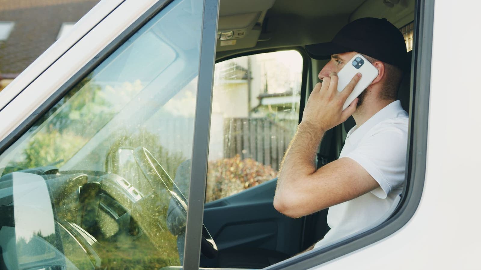 A man in a cap sits in the driver's seat of a white van talking on a phone