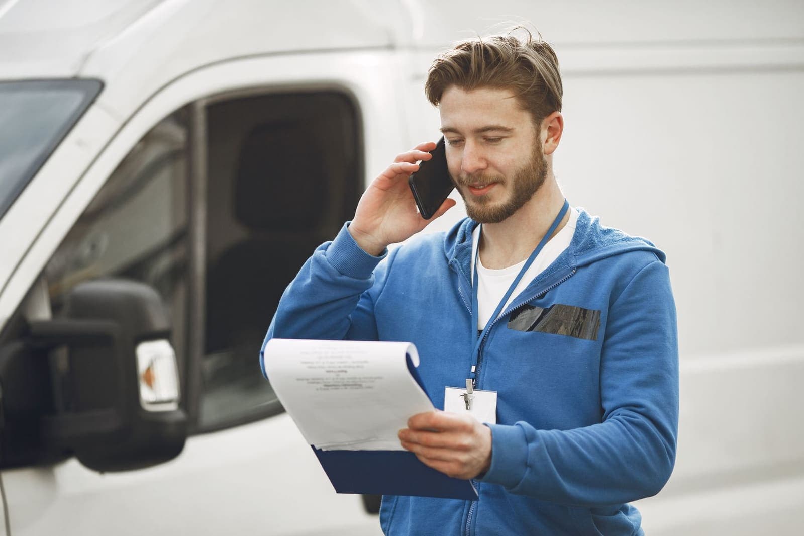 A tradesman in a blue hoodie holds a clipboard and talks on a phone beside a white work van