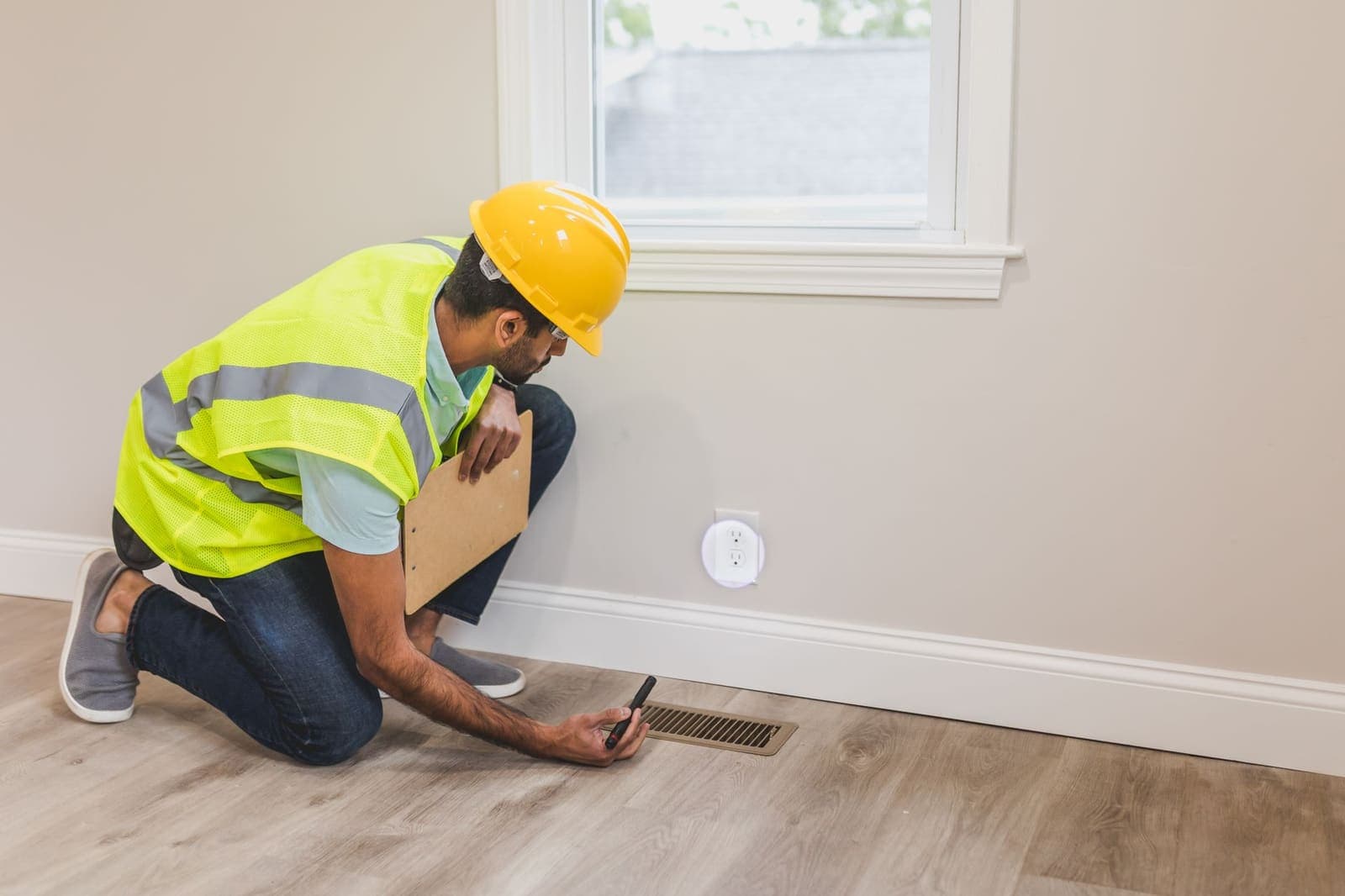 A contractor in a hard hat and high-visibility vest kneels by a floor vent with a flashlight and a clipboard