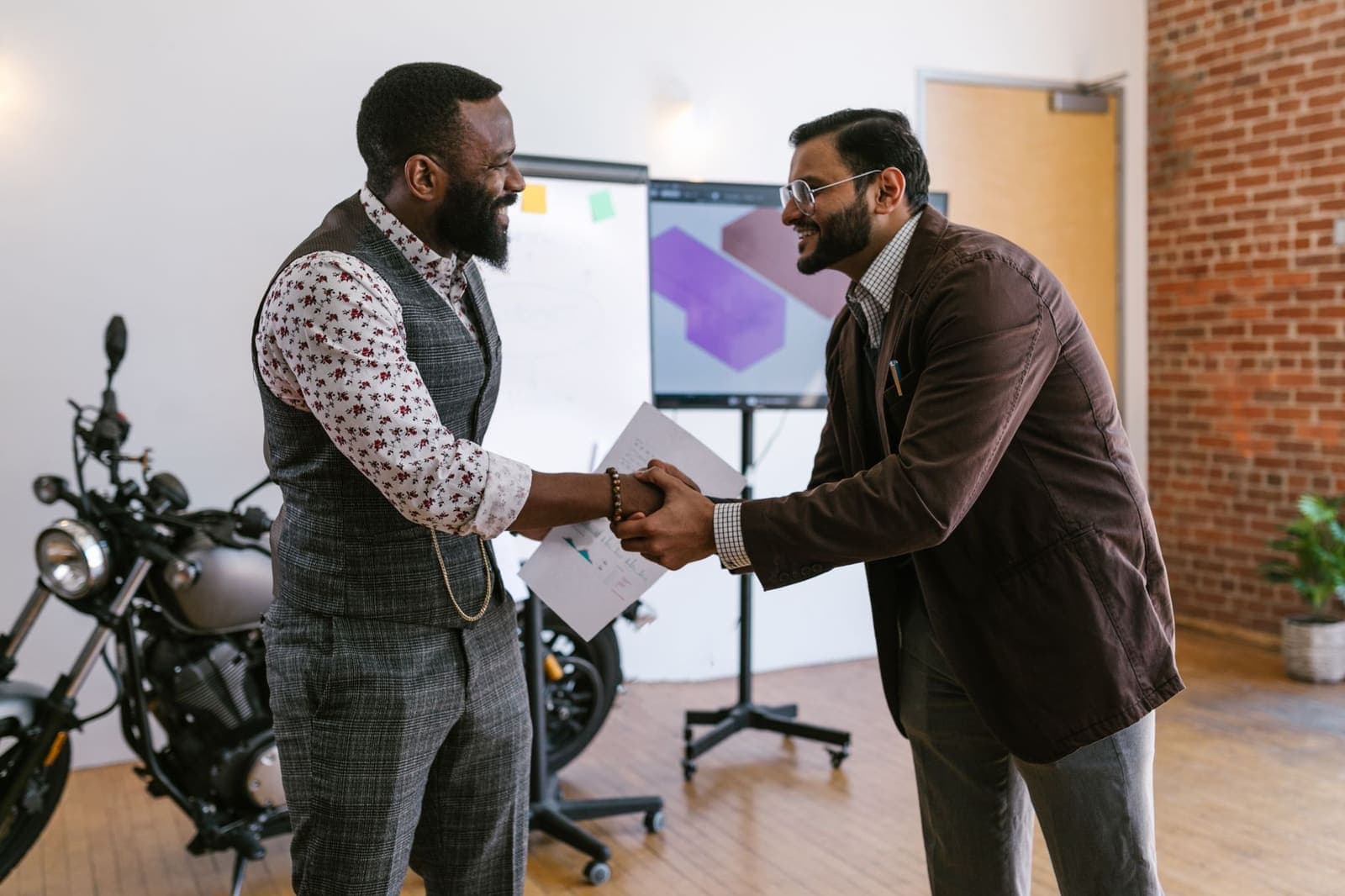 Two men shaking hands in a brick-walled workshop office with a motorcycle and a whiteboard behind them