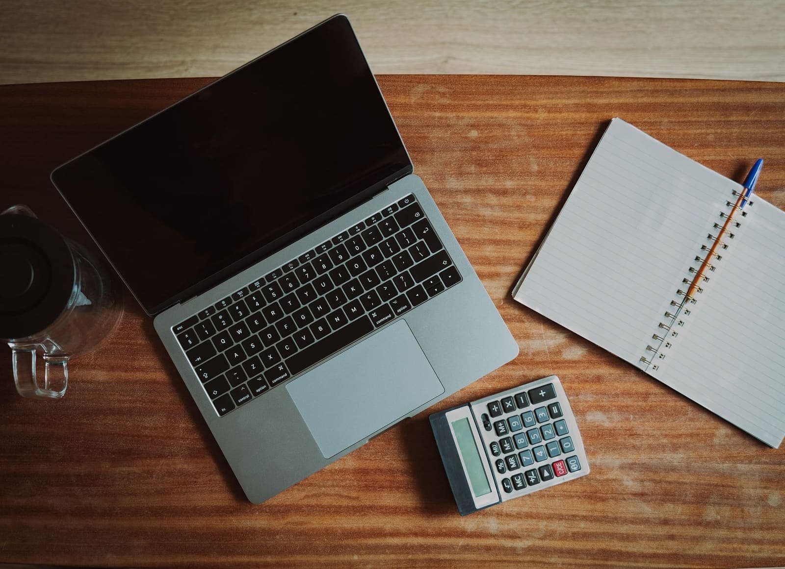 Overhead view of a laptop, a calculator and an open spiral notebook with a pen on a wooden desk