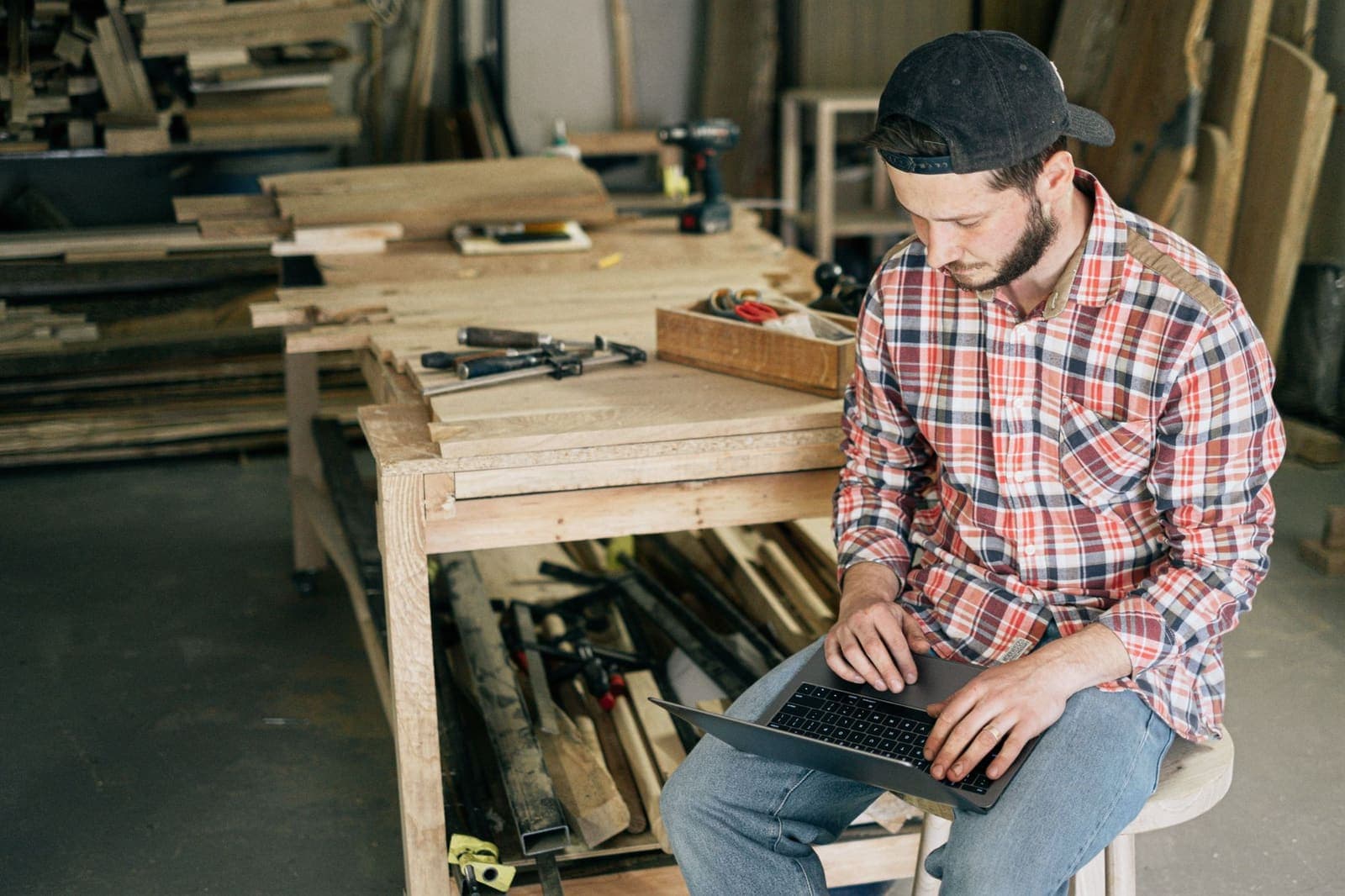 A tradesman in a plaid shirt and cap working on a laptop on a stool in a wood workshop, with clamps and boards on the bench behind him